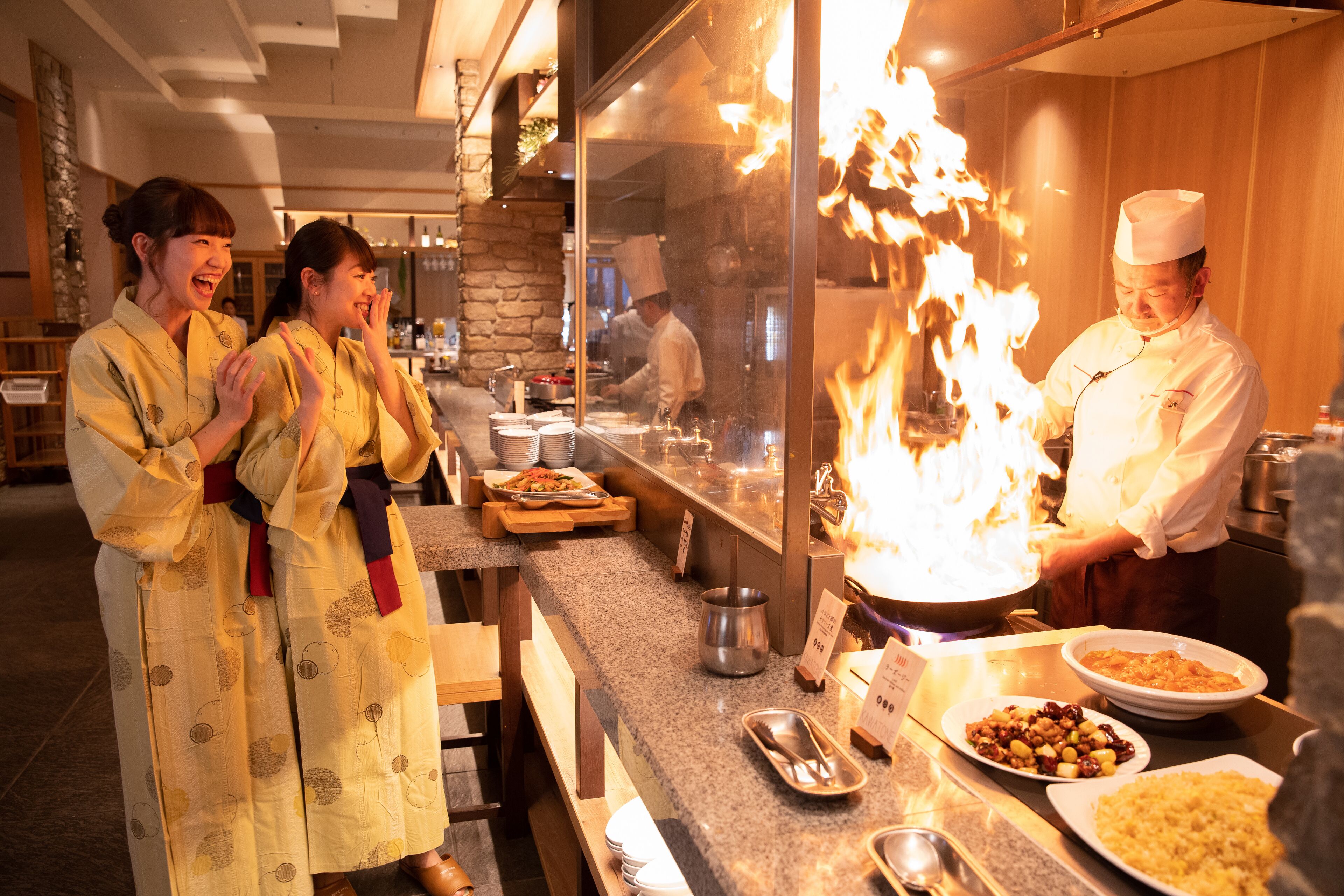 Restaurante com um chef apresentando um ato de fogo, um balcão com pratos e uma área de bar.