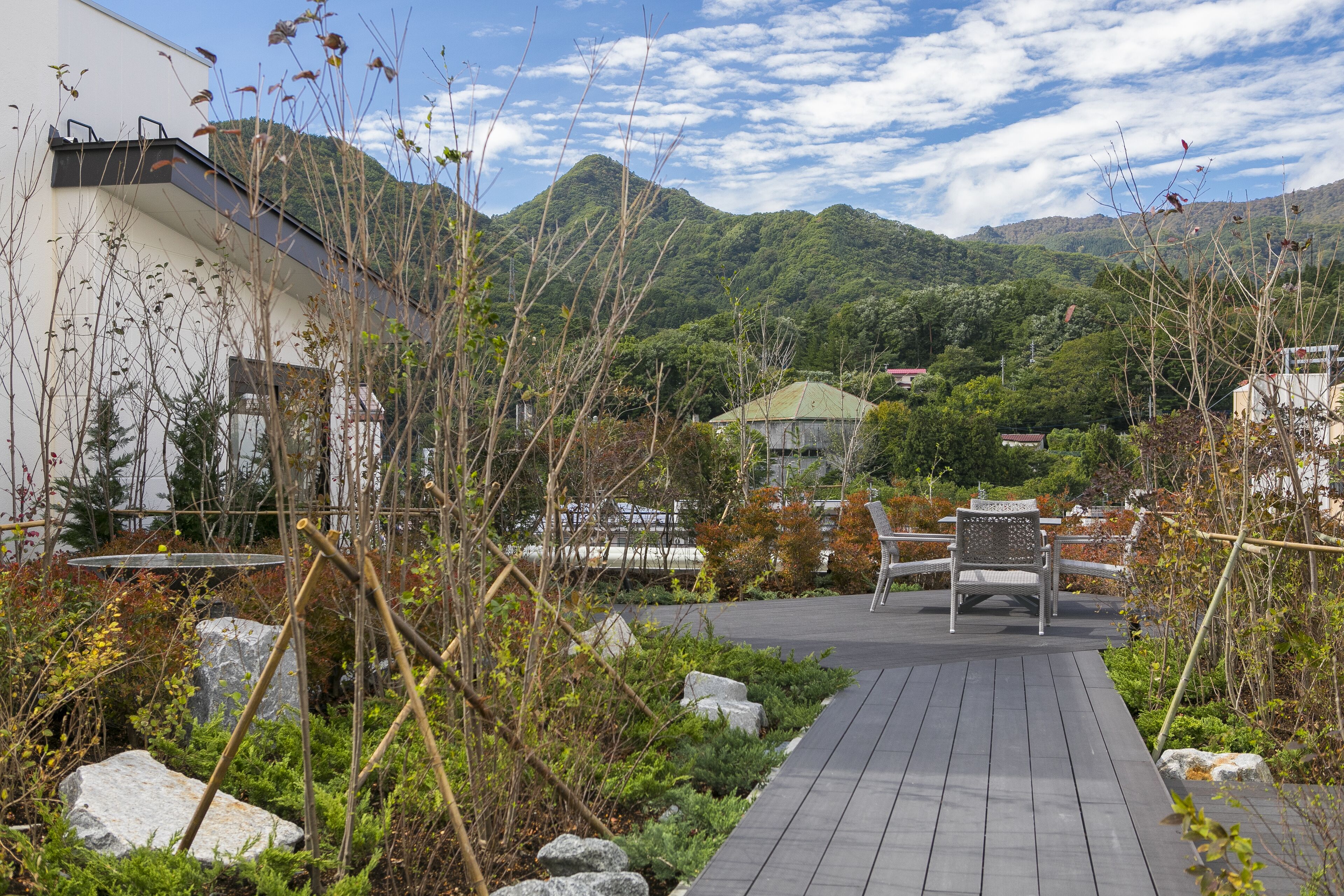 Une terrasse en bois avec une chaise, entourée de rochers et de plantes, qui mène à un bâtiment doté d’un toit végétalisé.