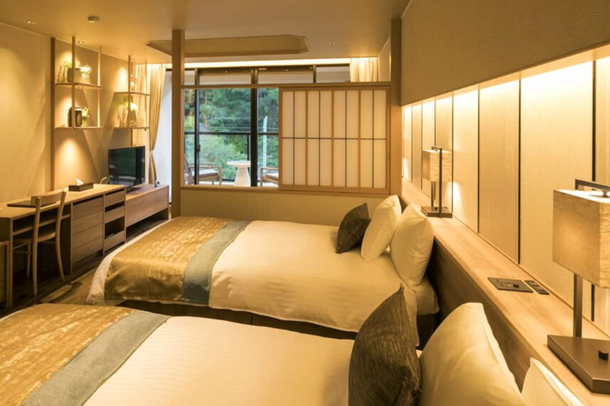 A hotel room with two beds, a desk, a TV, and a window with a view of greenery.