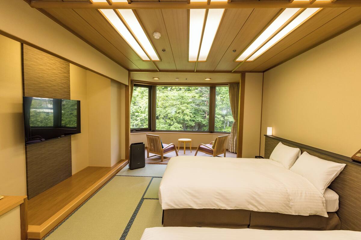 A hotel room with a bed, a TV mounted on the wall, a small table, two chairs, and large windows offering a view of greenery.