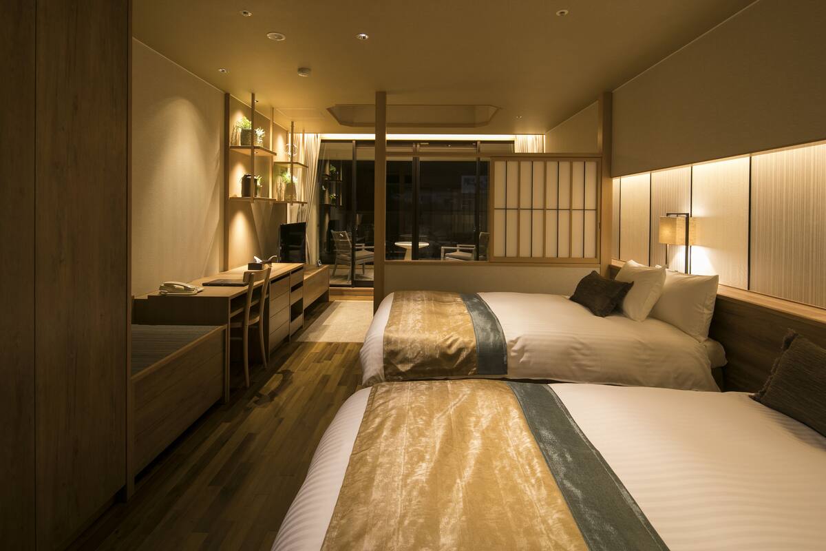 A hotel room with two beds, a desk, a chair, and a small table.