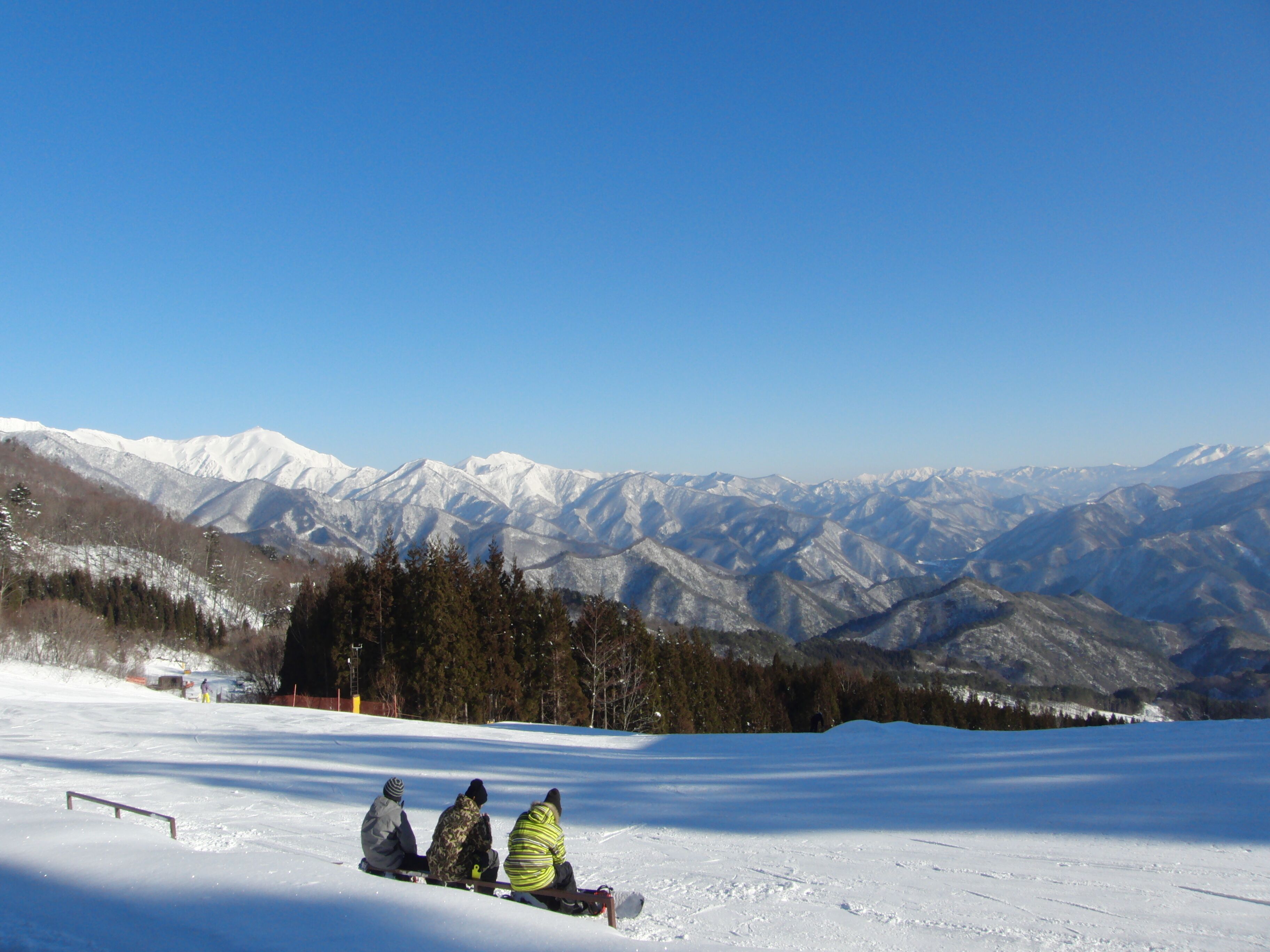Un paysage enneigé avec trois personnes assises sur une luge, un ciel bleu dégagé et des montagnes enneigées en arrière-plan.