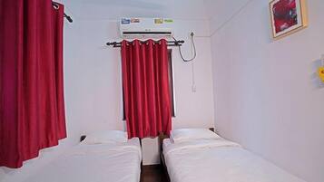 A room with two single beds, red curtains, an air conditioning unit, and a small painting on the wall.