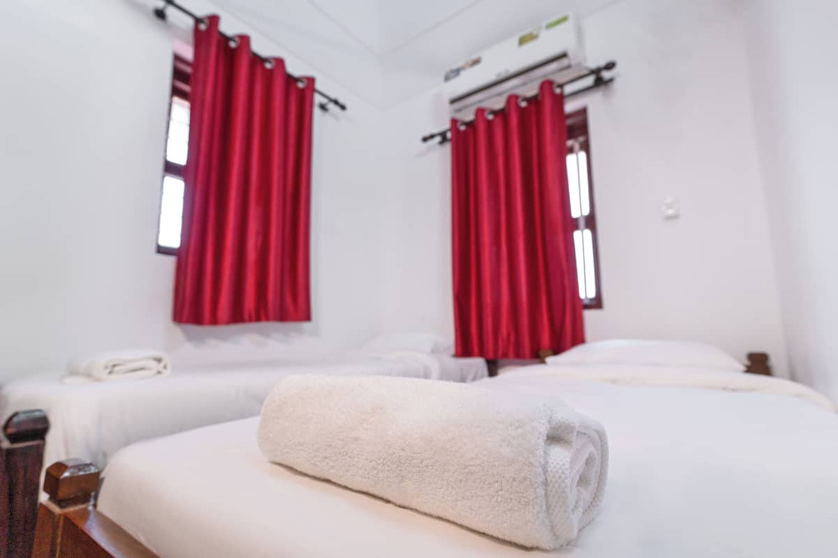 A hotel room with two beds, red curtains, white bedding, and a rolled towel on one bed.