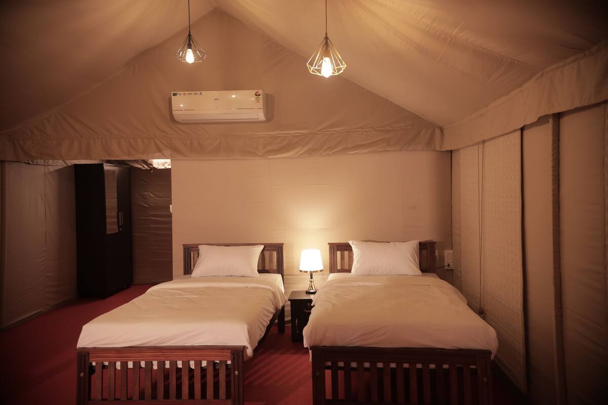 A room with two beds, a nightstand with a lamp, an air conditioner, and a hanging light fixture.