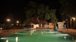 A nighttime pool area with a clear pool, surrounding trees, and a building in the background. - Anamala Serenity Homestay (Talappilly)