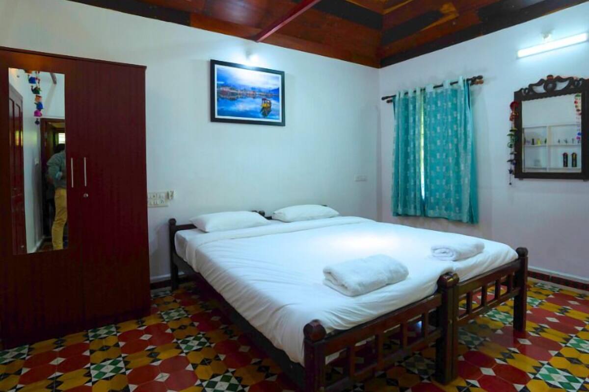A bedroom with a wooden bed, a colorful geometric floor pattern, a wooden wardrobe, a painting on the wall, and a window with curtains.