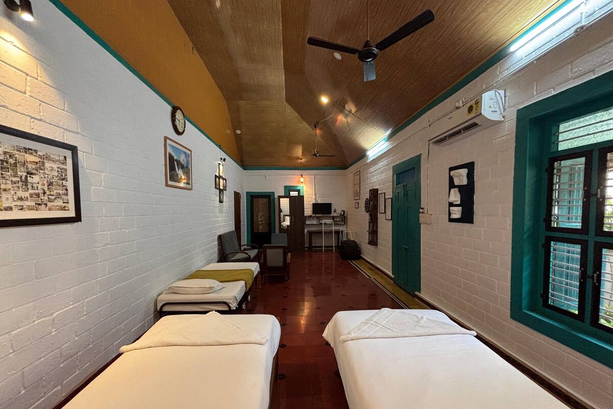 A corridor with four single beds lined up against a white brick wall, a ceiling fan, and a window with bars.