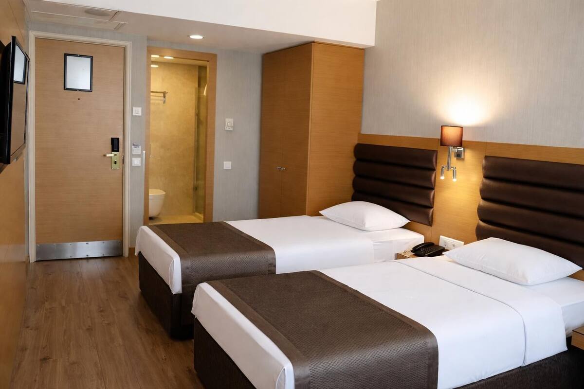 A hotel room with two beds, a bedside table with a lamp, a wooden wardrobe, and a bathroom visible through an open door.