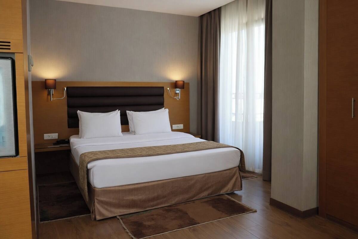 A modern hotel room with a large bed, wooden headboard, two bedside lamps, and a view of the outdoors through sheer curtains.