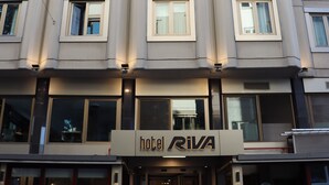 Un hôtel à plusieurs étages, doté d’une façade moderne, de grandes fenêtres et d’une enseigne bien visible, « Hôtel RIVA », à l’entrée.