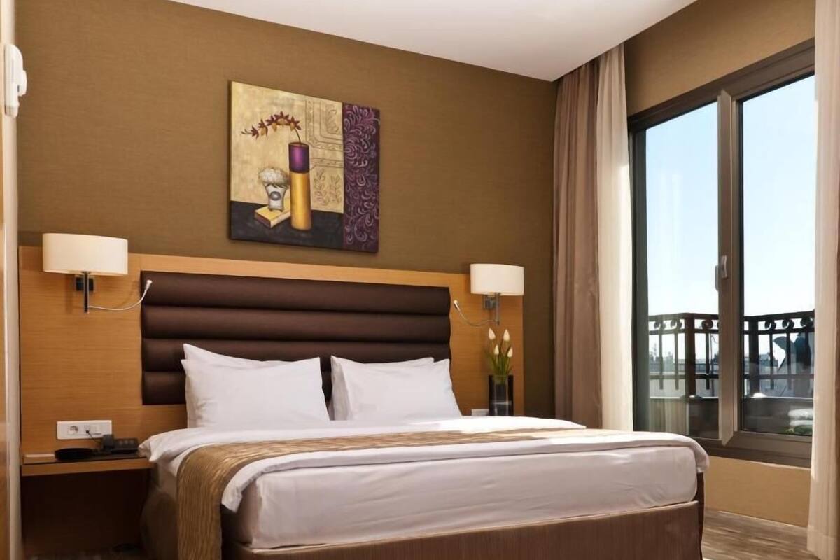 A hotel room with a large bed, two bedside tables, a wall-mounted artwork, a window with a view, and a balcony.