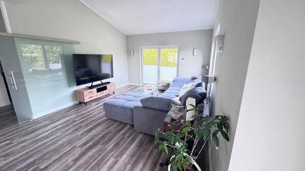 Ein modernes Wohnzimmer mit einem großen Flachbildfernseher, einem grauen Ecksofa und einem Glastisch.