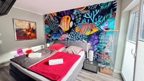 Um quarto com cama, mesa de cabeceira, porta de vidro e um mural colorido na parede.