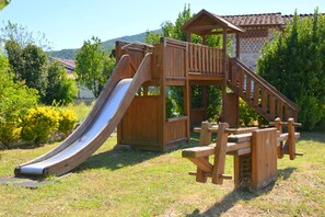 Children can play safely on the playground's slide and climbing area amidst lush green surroundings.