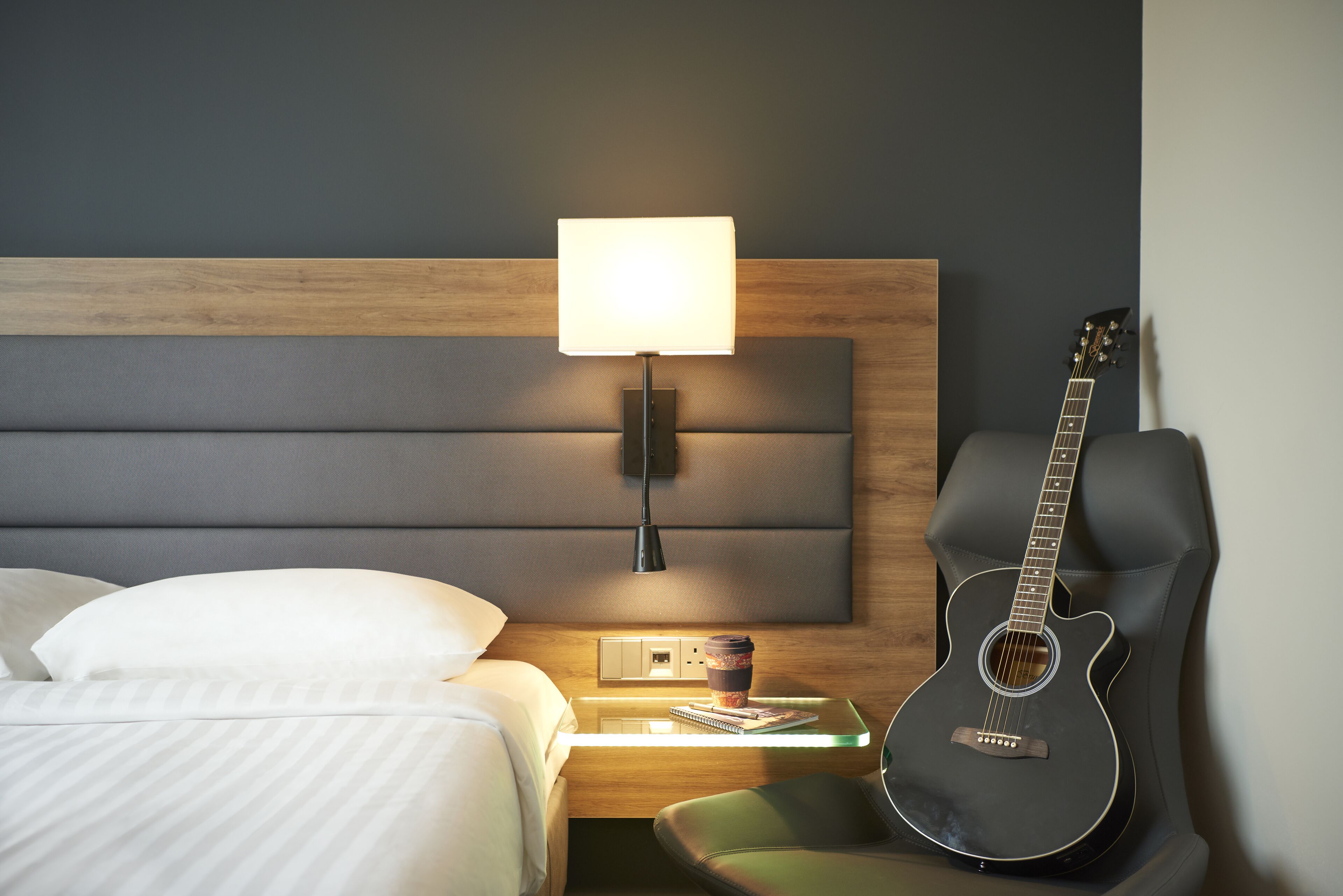 A hotel room with a bed, a nightstand with a lamp, a guitar, and a cup of coffee.