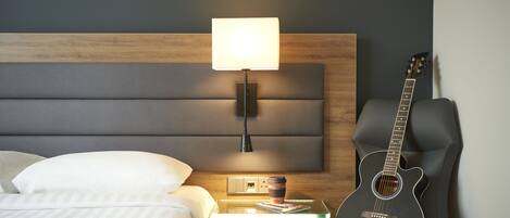 A hotel room with a bed, a nightstand with a lamp, a guitar, and a cup of coffee.