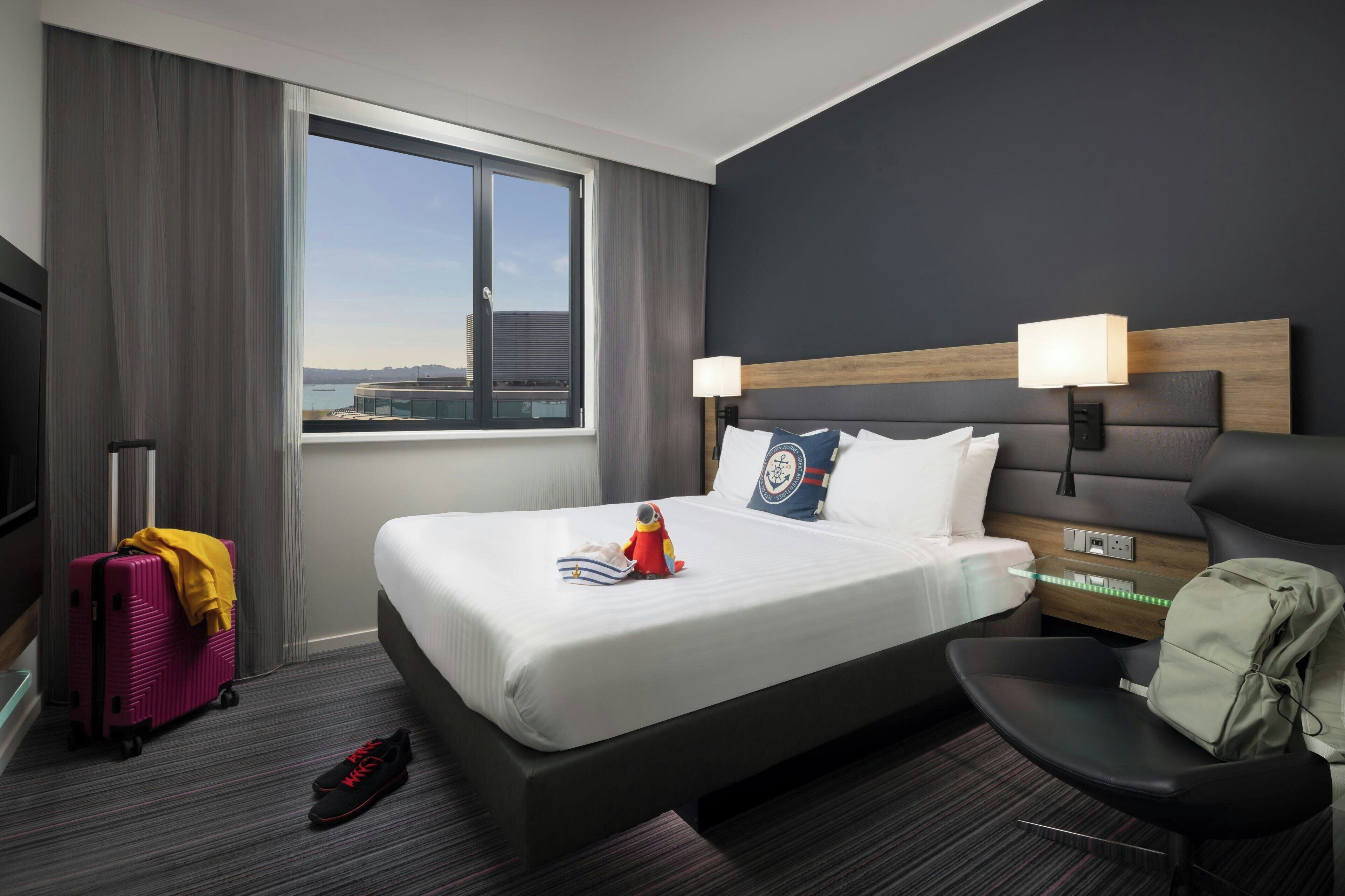 A hotel room with a large bed, a desk, a chair, a suitcase, and a view of a cityscape.