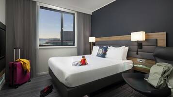Habitación de hotel con una cama grande, un escritorio, una silla, una maleta y vistas a un paisaje urbano.