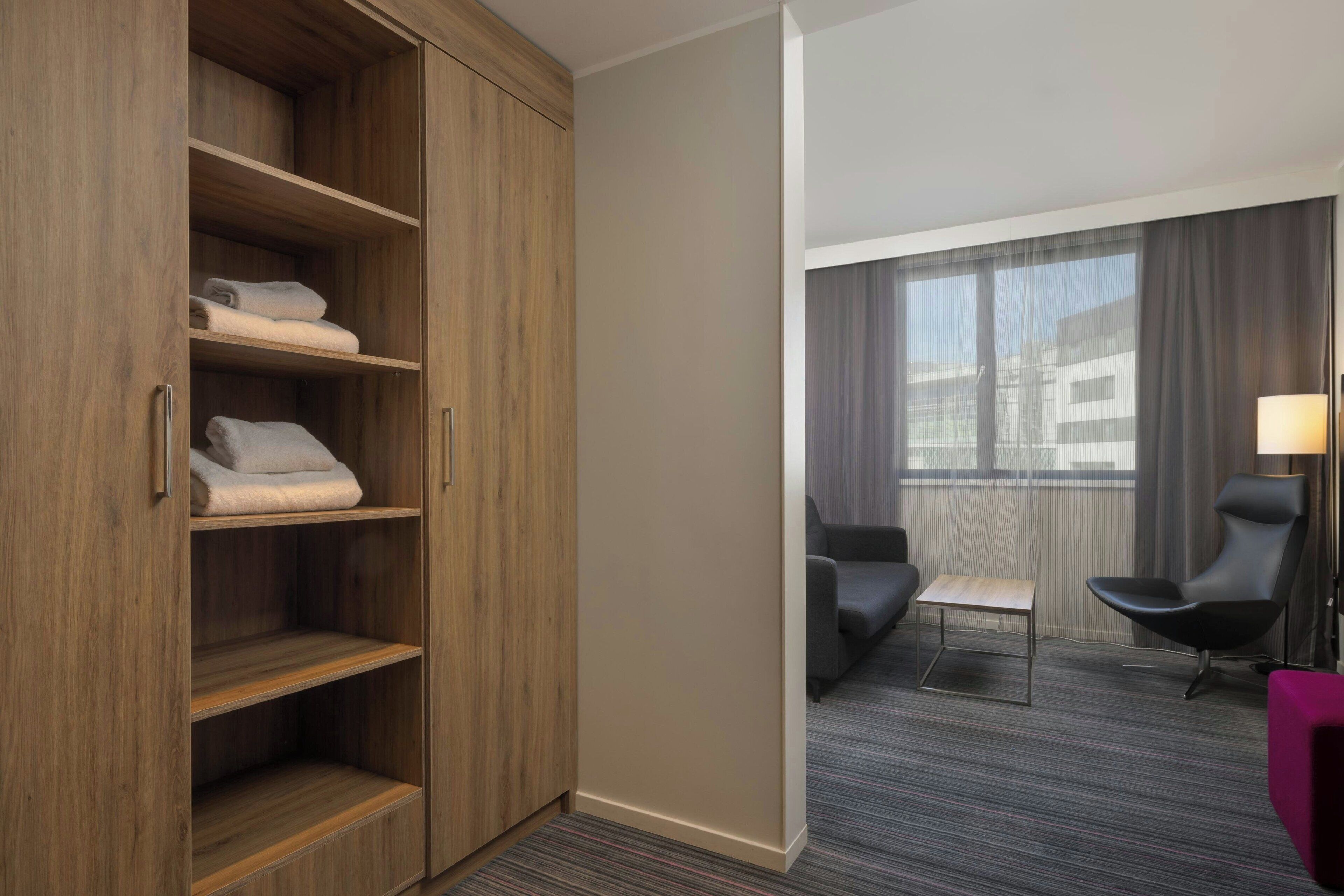 A hotel room with a wooden wardrobe, a seating area with a sofa and armchair, and a small table.
