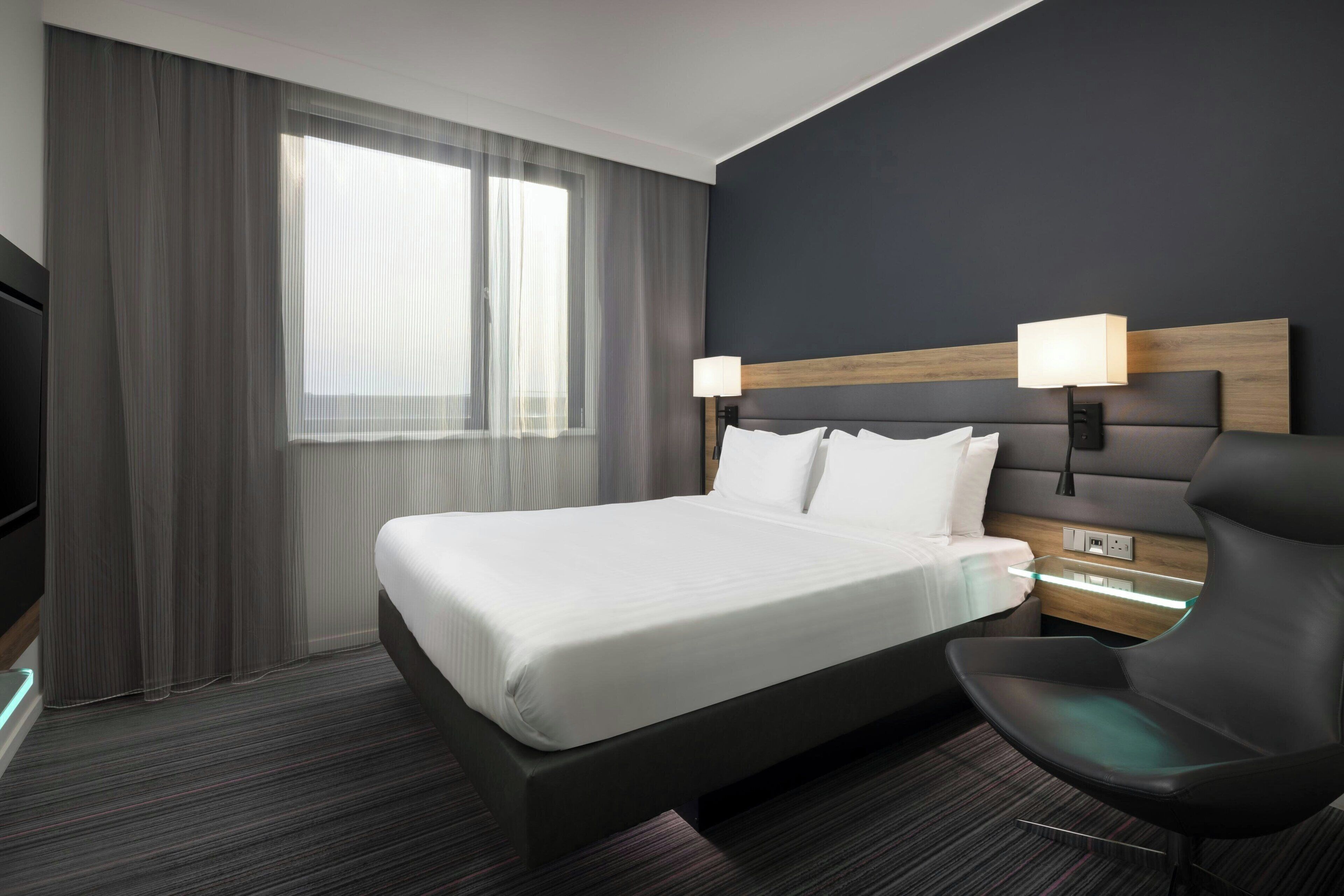 Una moderna camera d'hotel con un grande letto, un comodino con lampada, una poltrona e una televisione.