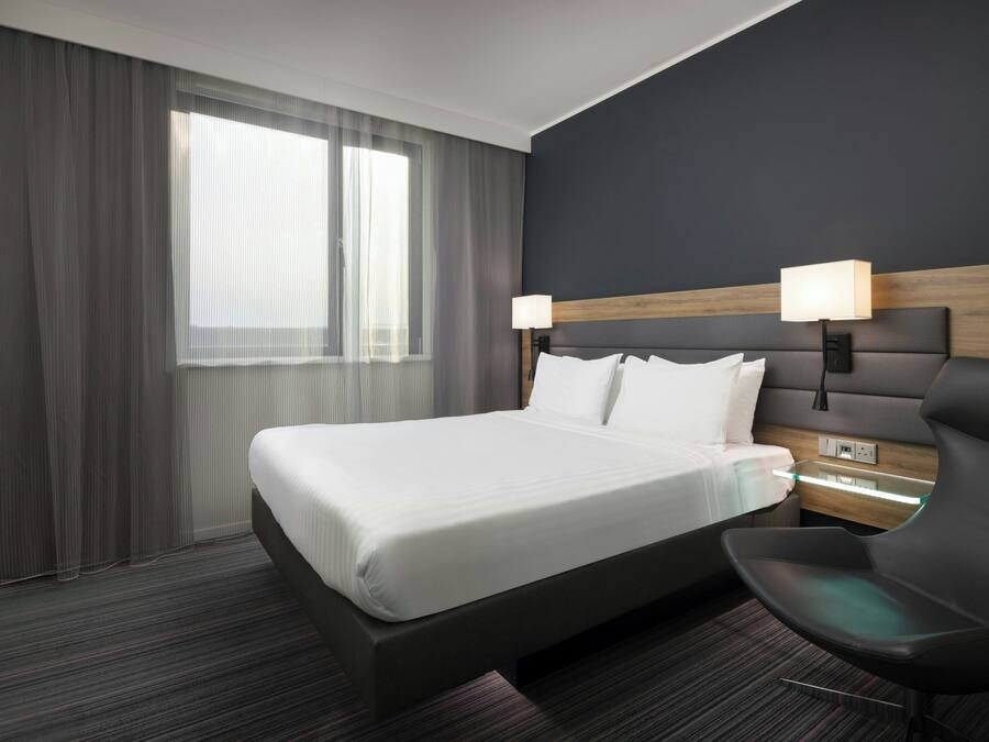 Una moderna camera d'hotel con un grande letto, un comodino con lampada, una poltrona e una televisione.