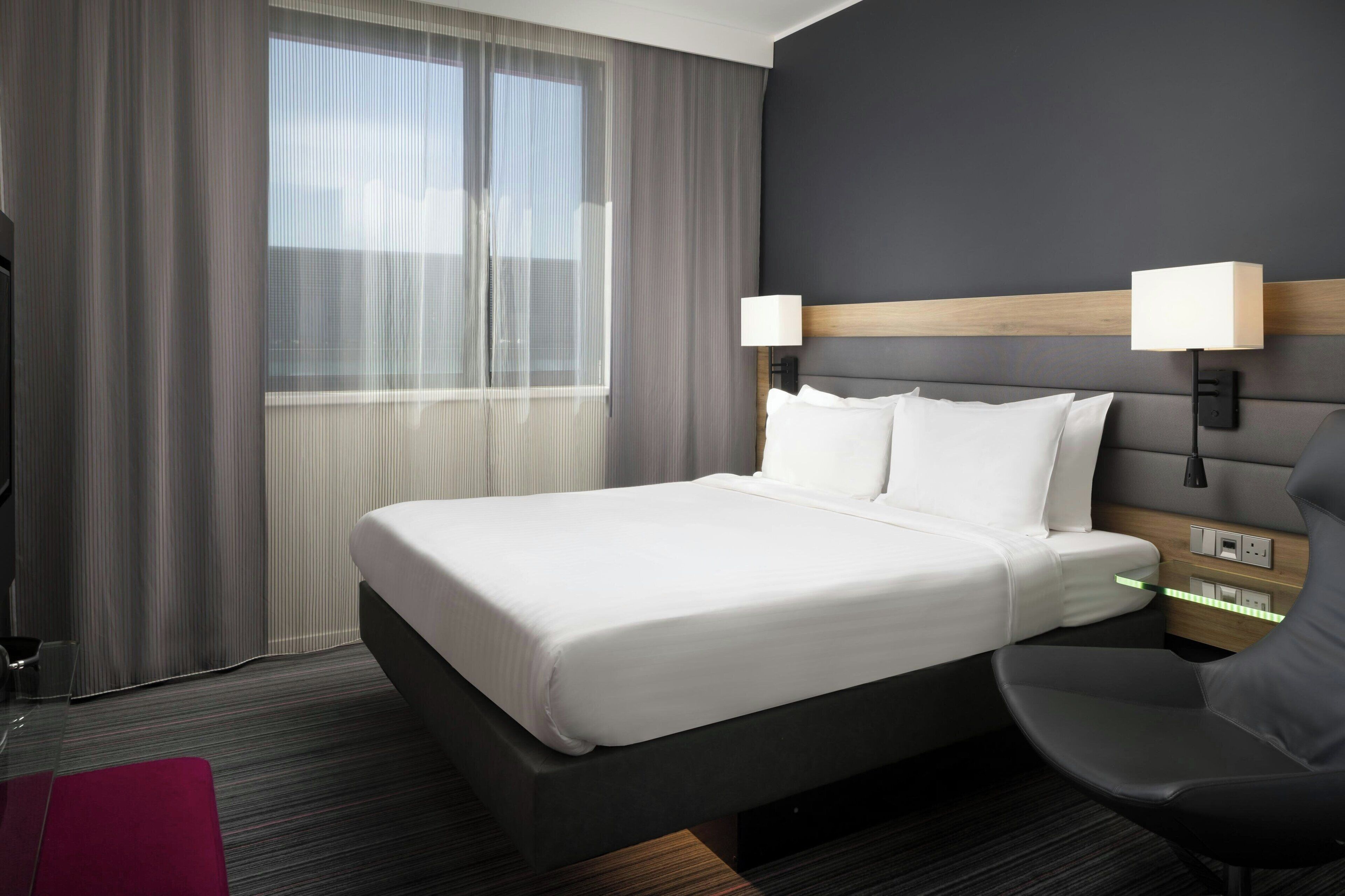 A hotel room with a large bed, two bedside lamps, a chair, a desk, and a window with curtains.
