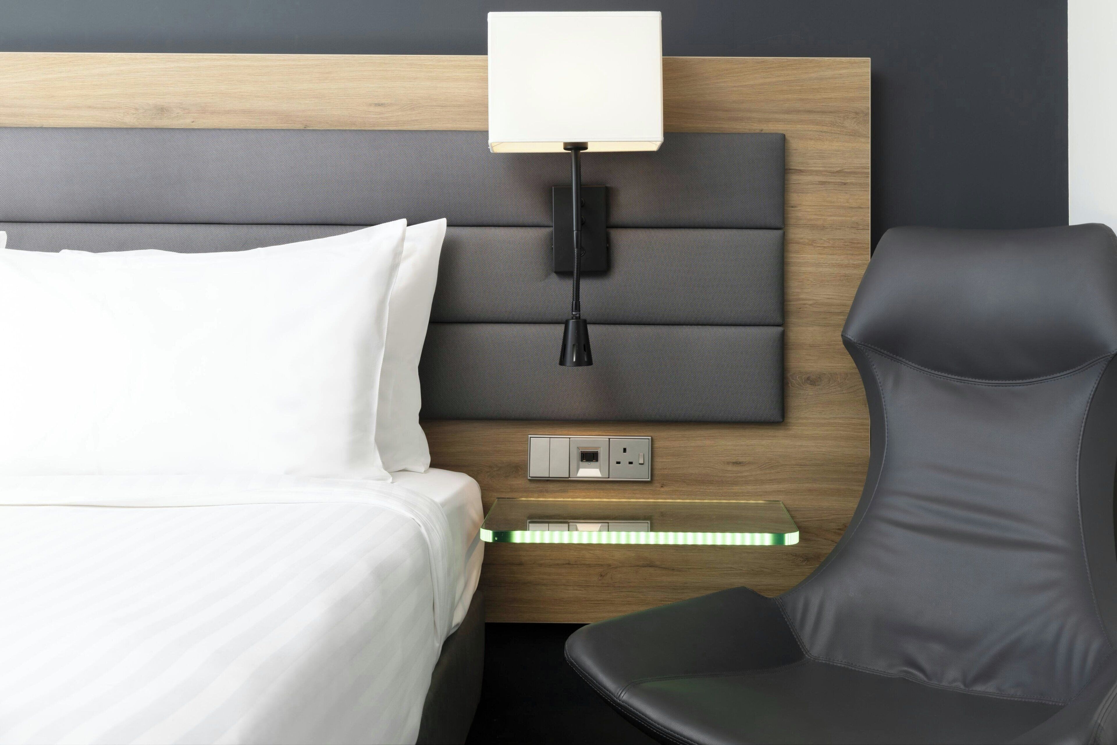 A modern hotel room with a wooden headboard, a bedside table with a lamp, a chair, and a power outlet.