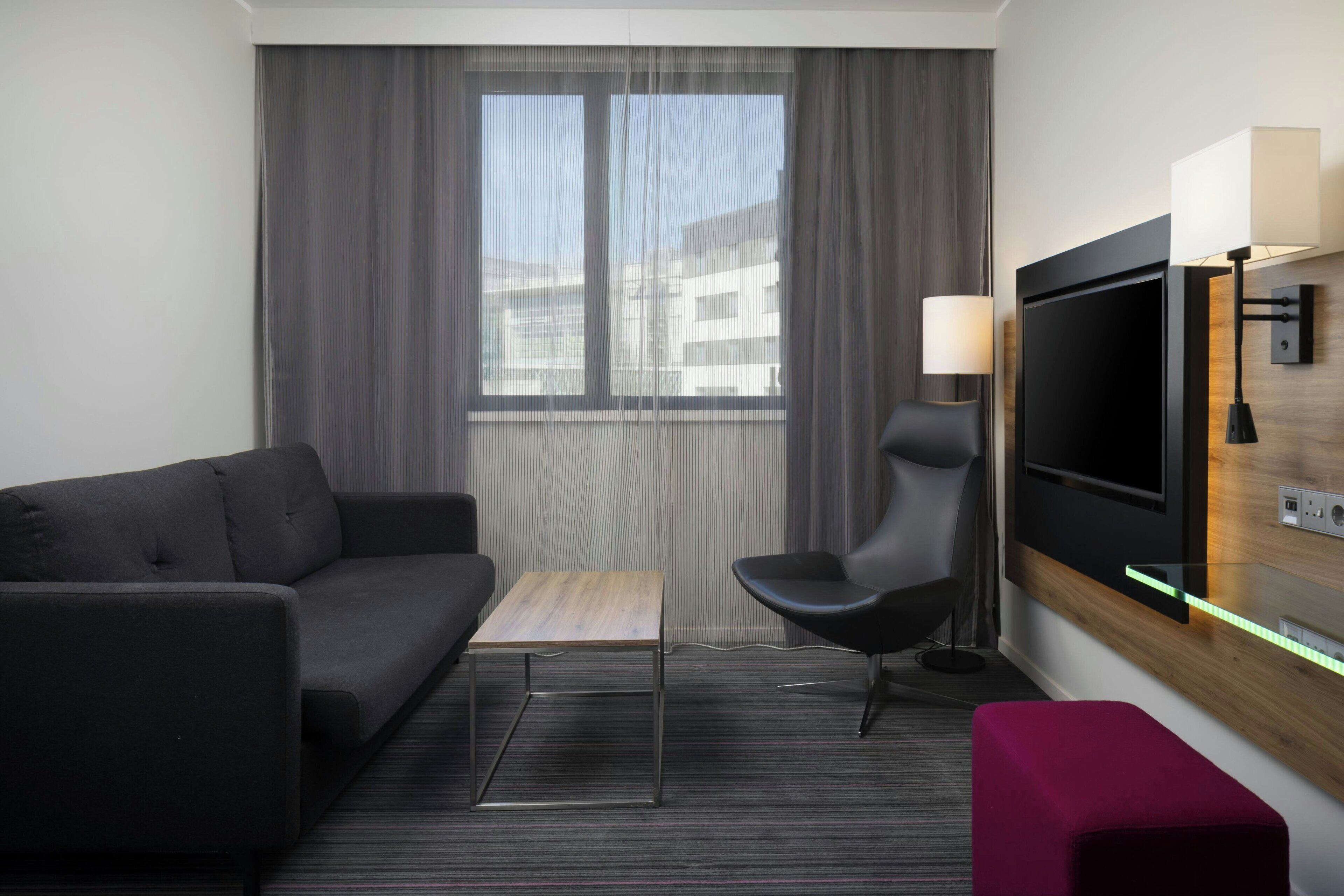 A modern hotel room with a dark grey sofa, a wooden coffee table, a black armchair, a flat-screen TV mounted on the wall, and a small wooden side table.