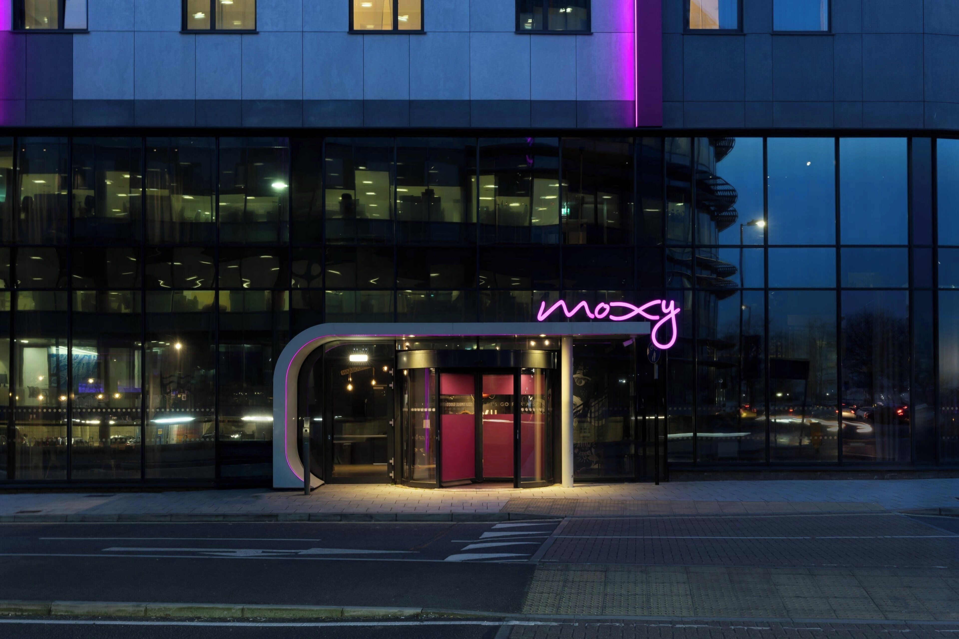 Un edificio moderno con fachada de vidrio y un letrero de neón que dice "Moxy".