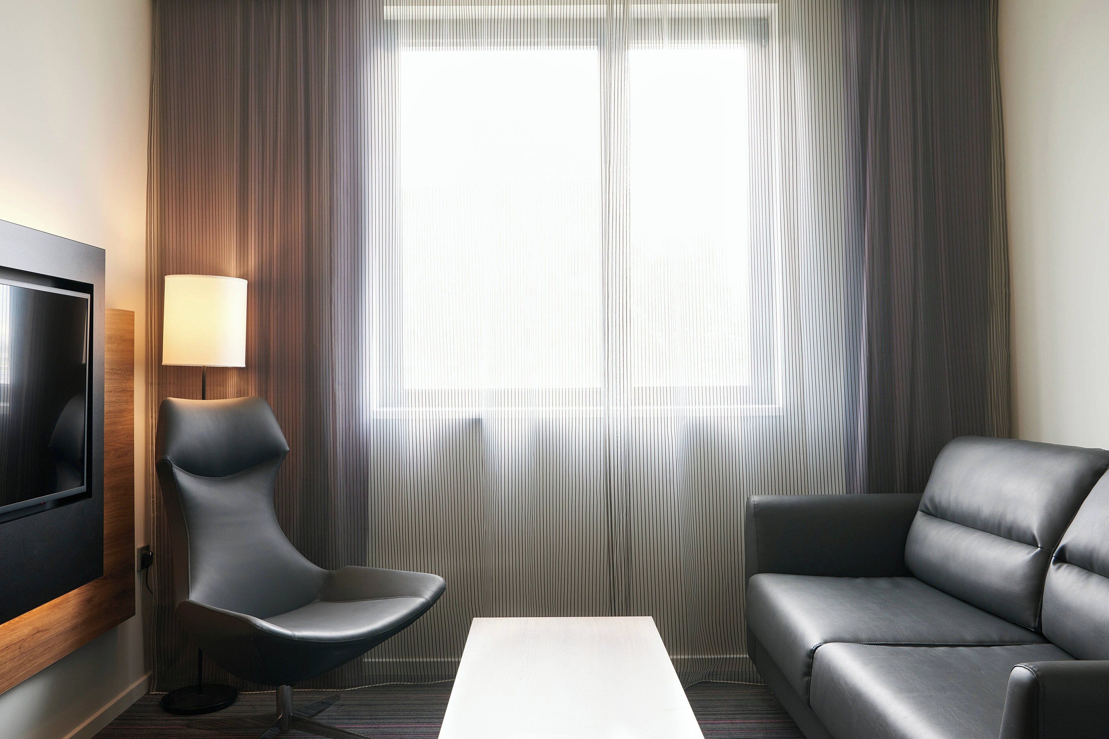 A modern hotel room with a flat-screen TV, a grey sofa, a black armchair, and a white coffee table.