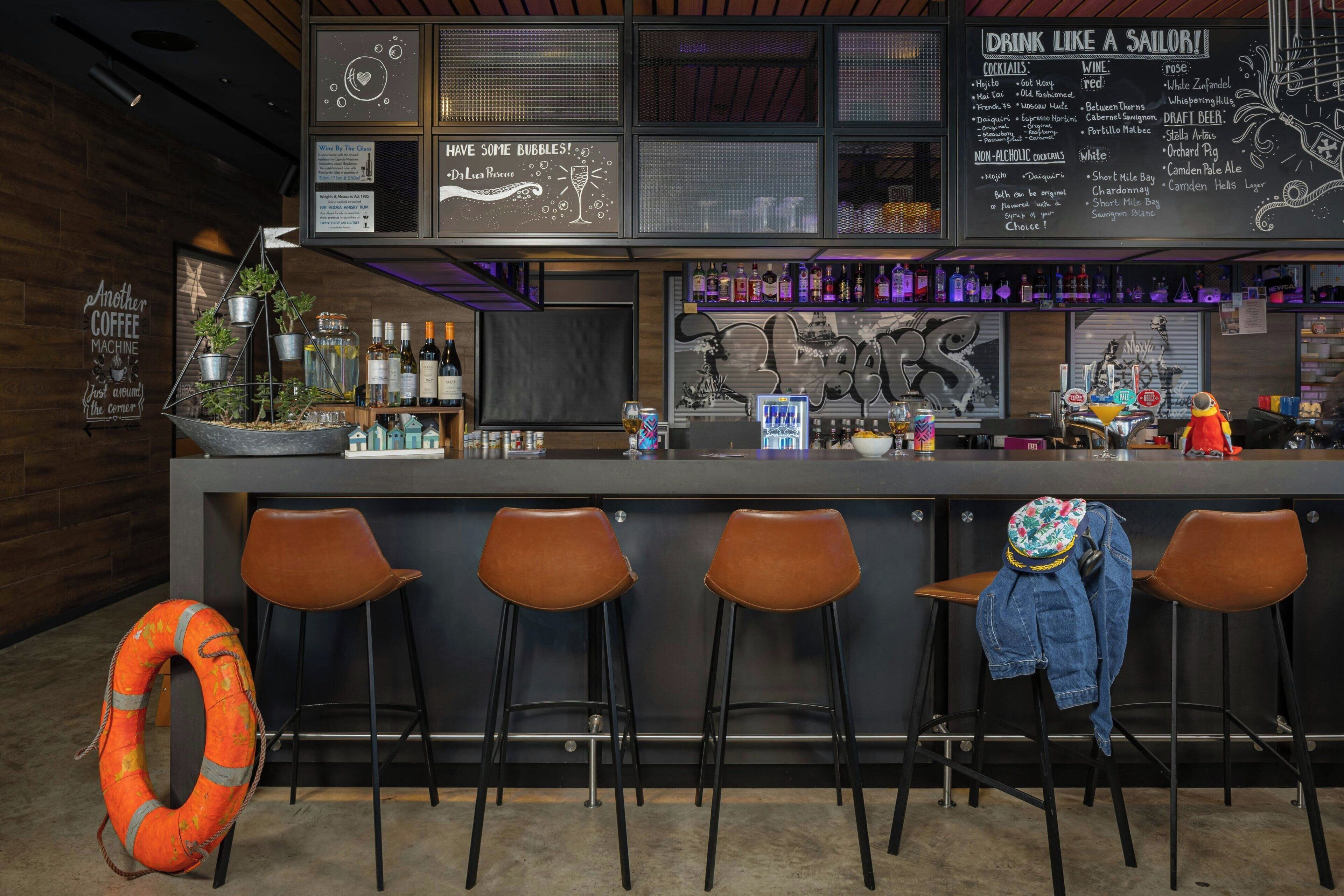 A bar area with stools, a neon sign, and a variety of bottles and glasses on the shelves.