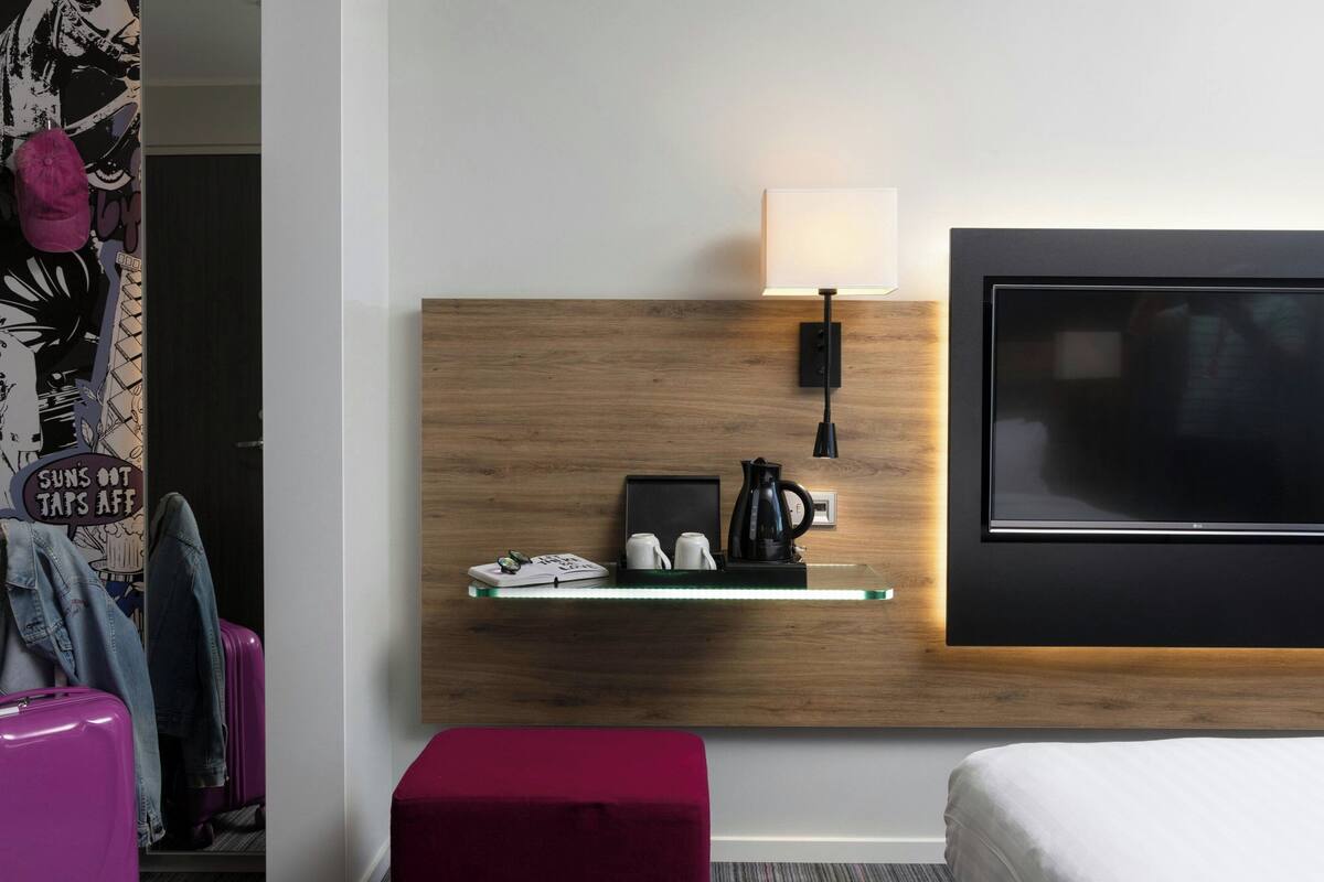 A modern hotel room with a wooden headboard, a flat-screen TV, a bedside table with a lamp, and a red ottoman.