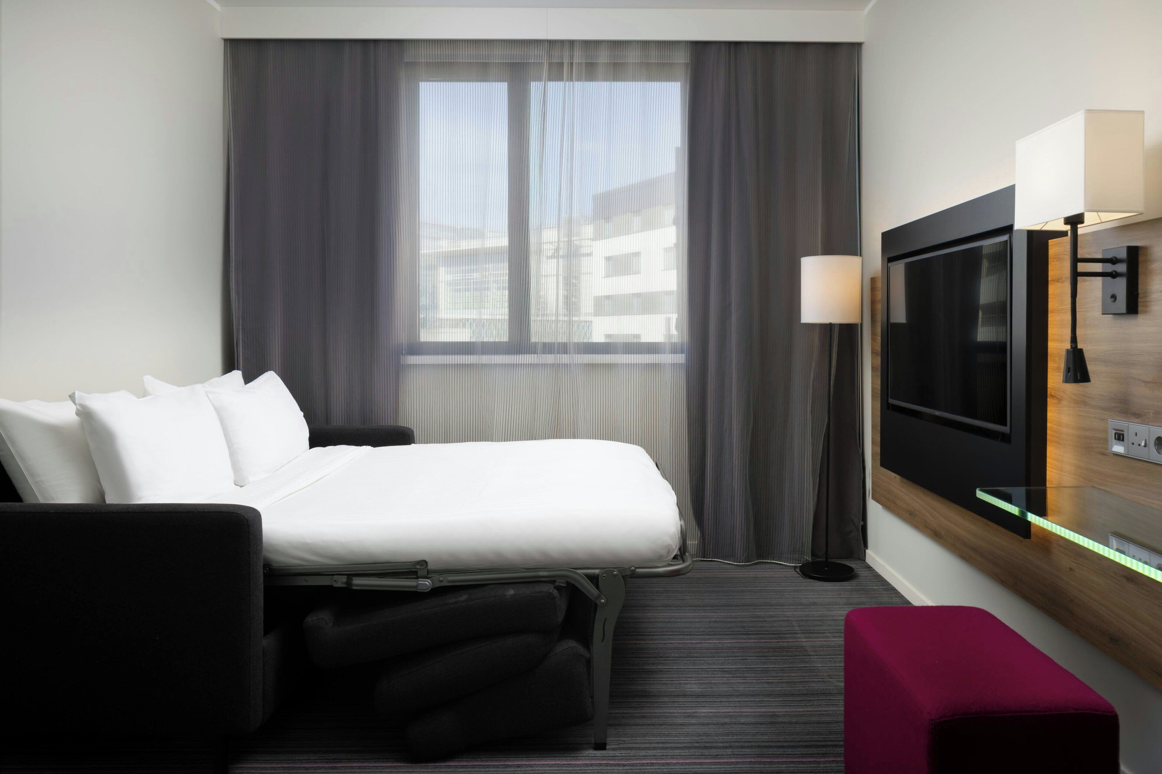 A hotel room with a bed, a TV, a desk with a lamp, a window with curtains, and a red bench.