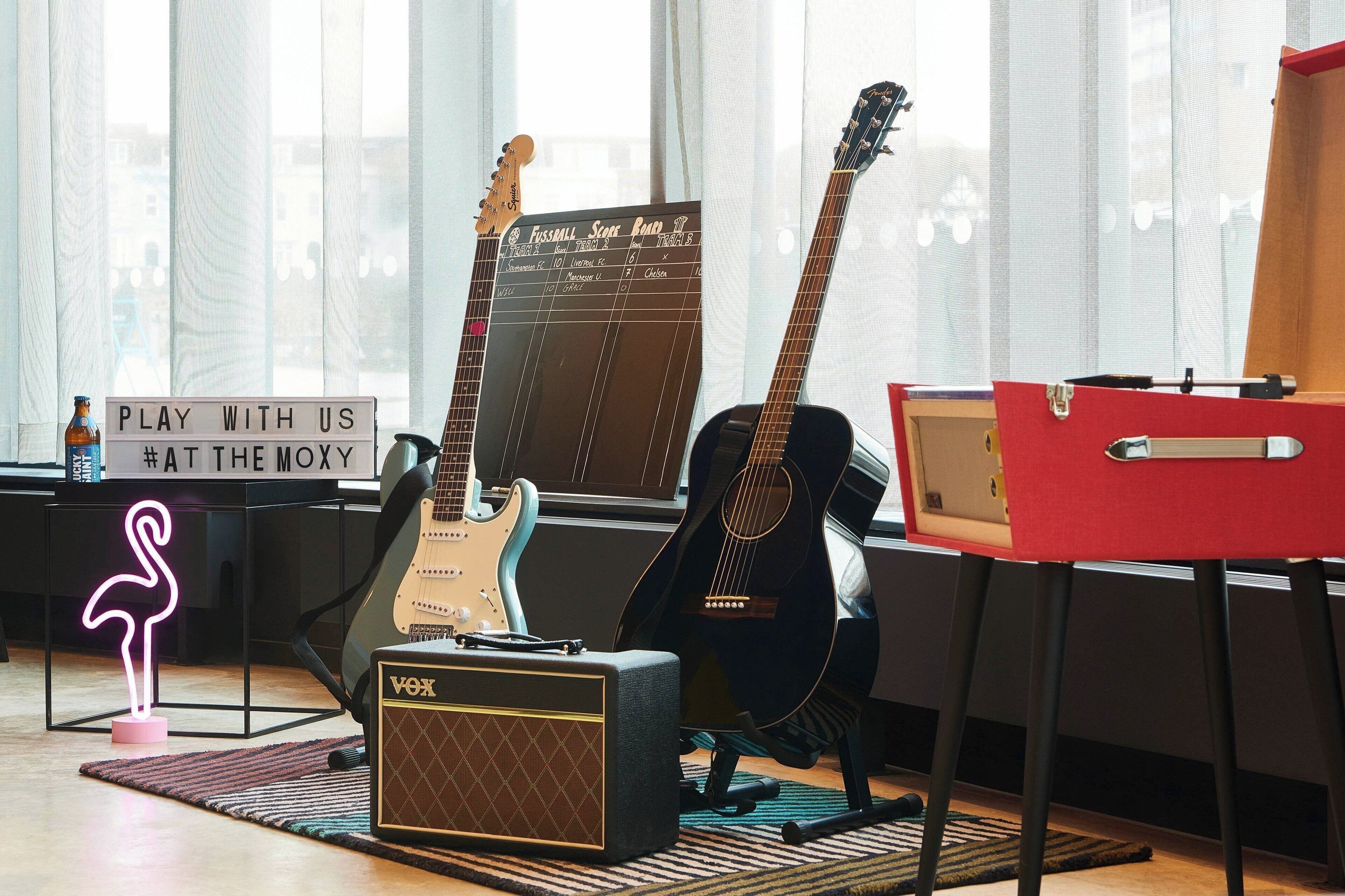 A room with a guitar, amplifier, and a sign that reads "PLAY WITH US #ATTHEMOXY".