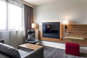 Habitación de hotel con televisor montado en la pared, un sofá, una silla, una mesita y un reposapiés rojo.