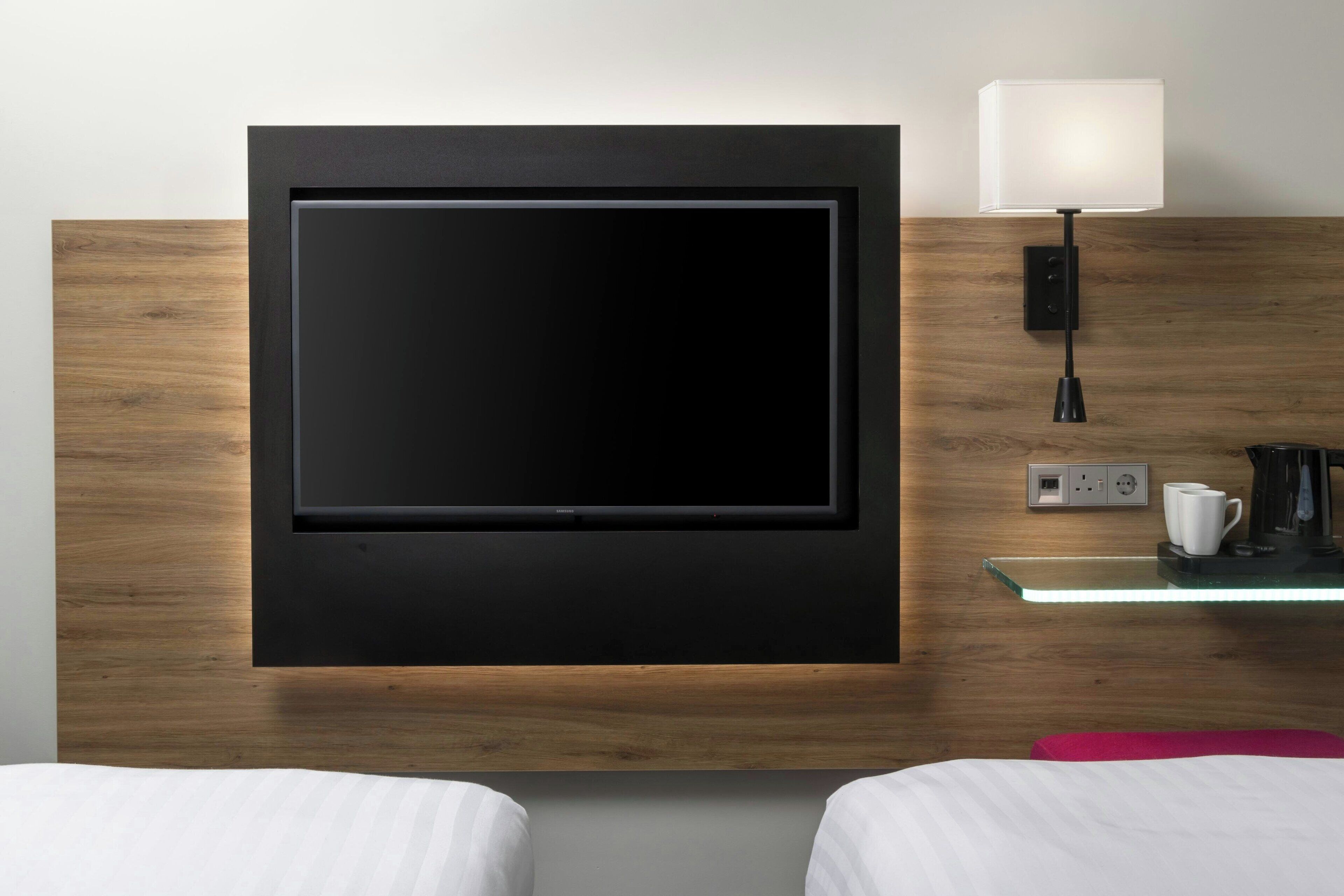 A modern hotel room with a large flat-screen TV mounted on a wooden wall, a bedside table with a lamp, and a coffee maker.
