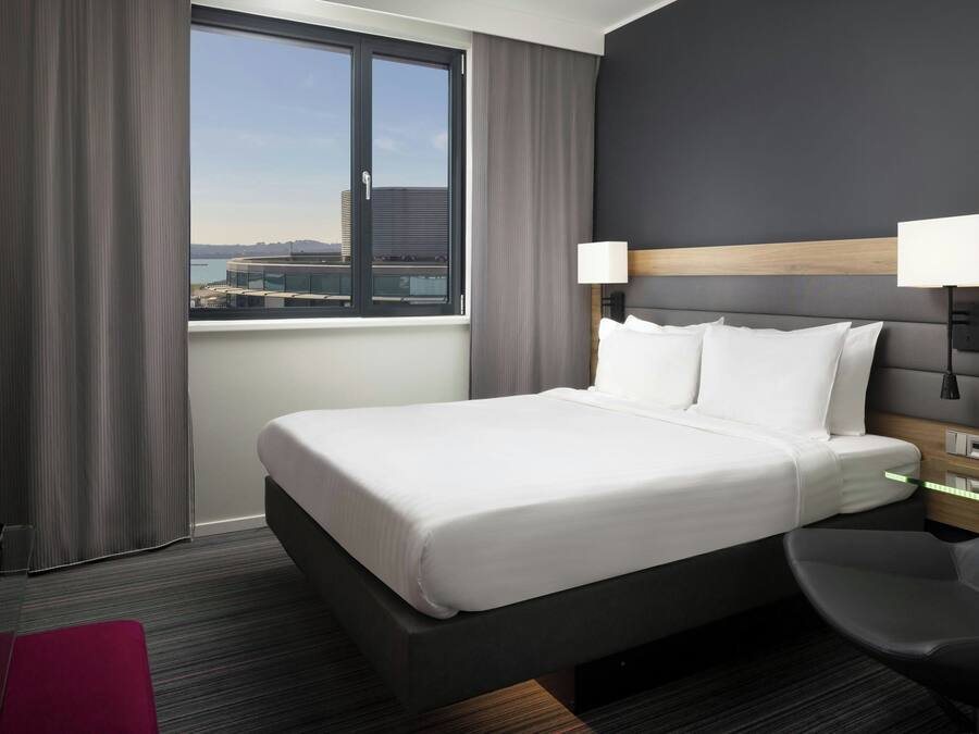 A hotel room with a large bed, two bedside lamps, a view of a cityscape, a desk, and a chair.