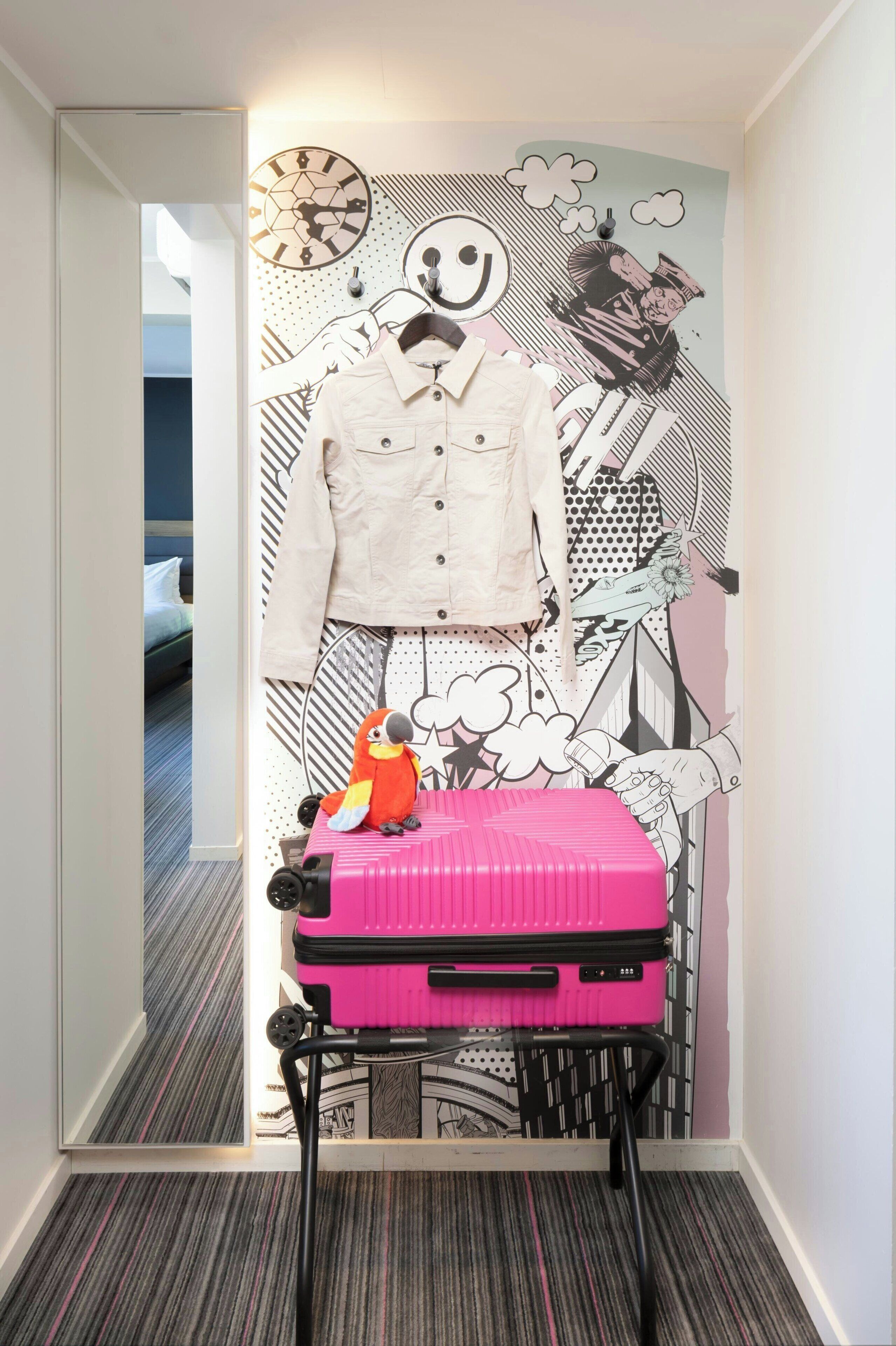 A hallway with a wall mural, a hanging jacket, a pink suitcase, and a toy parrot.