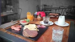 Una mesa de desayuno con diversos alimentos, como huevos, salchicha y una hamburguesa a la parrilla, además de bebidas y un jarrón con flores.