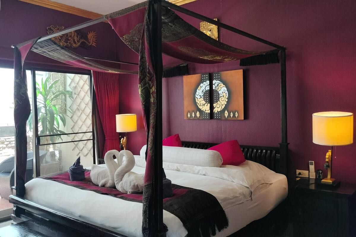 A four-poster bed with white linens, red pillows, and a red throw blanket. There are two wall-mounted lamps and a large painting above the bed.
