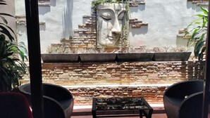 An outdoor seating area with wicker chairs and a table, a stone wall with a decorative face, and potted plants. - Le Prive Resort (Pattaya)