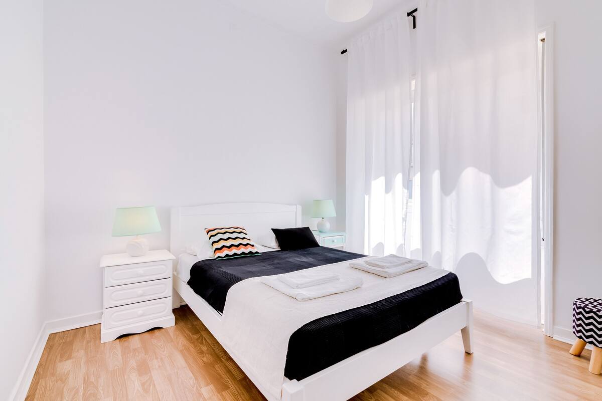 A modern bedroom with a white bed, black and white bedding, and a nightstand with a lamp.