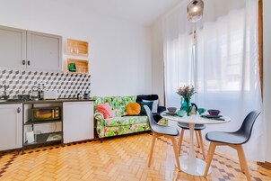 Cocina compacta con microondas, pileta y alacenas. Hay un comedor con mesa, sillas y un sofá estampado con motivos tropicales.