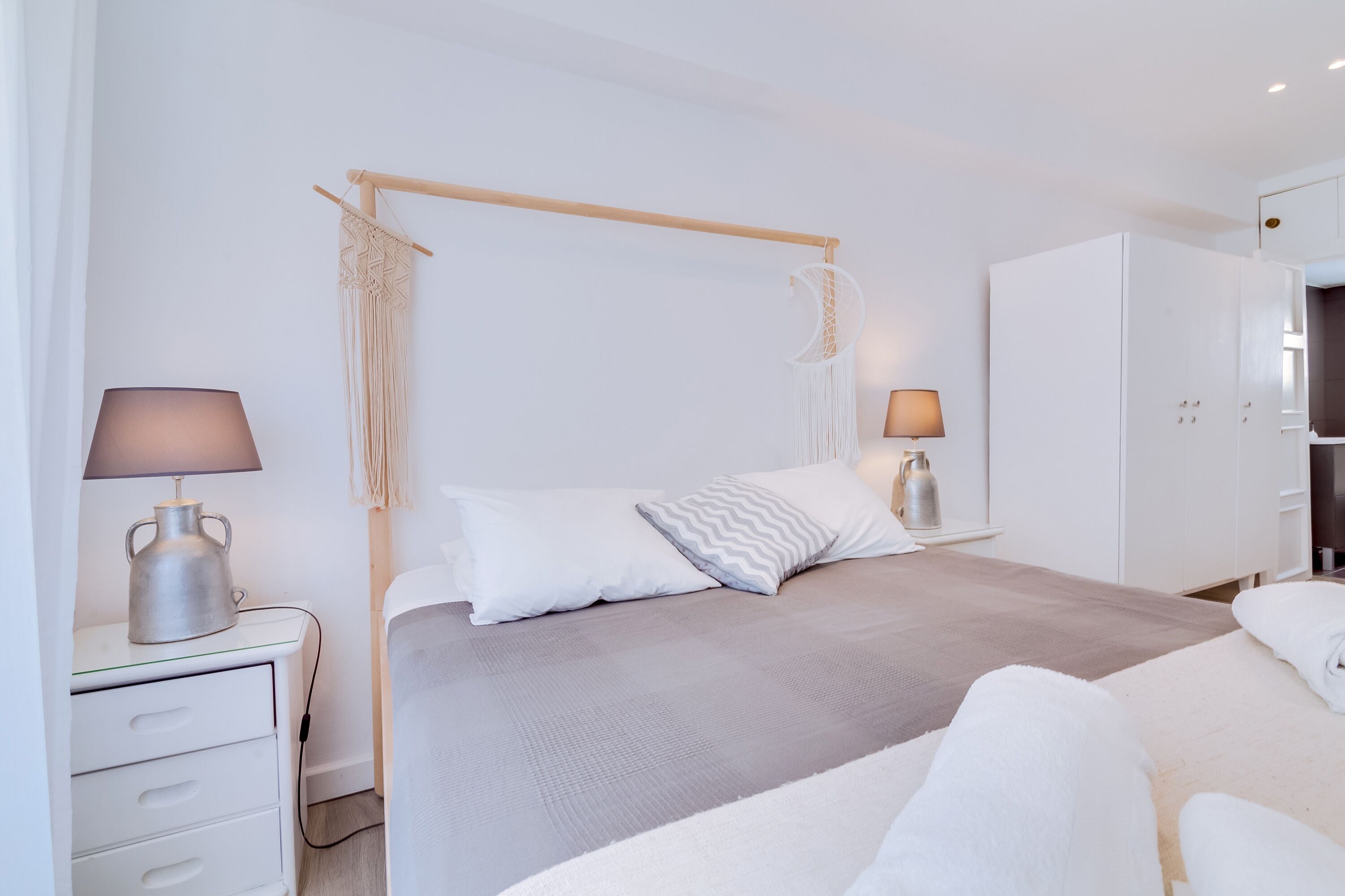 A modern bedroom with a four-poster bed, bedside tables with lamps, a wardrobe, and a macrame wall hanging.