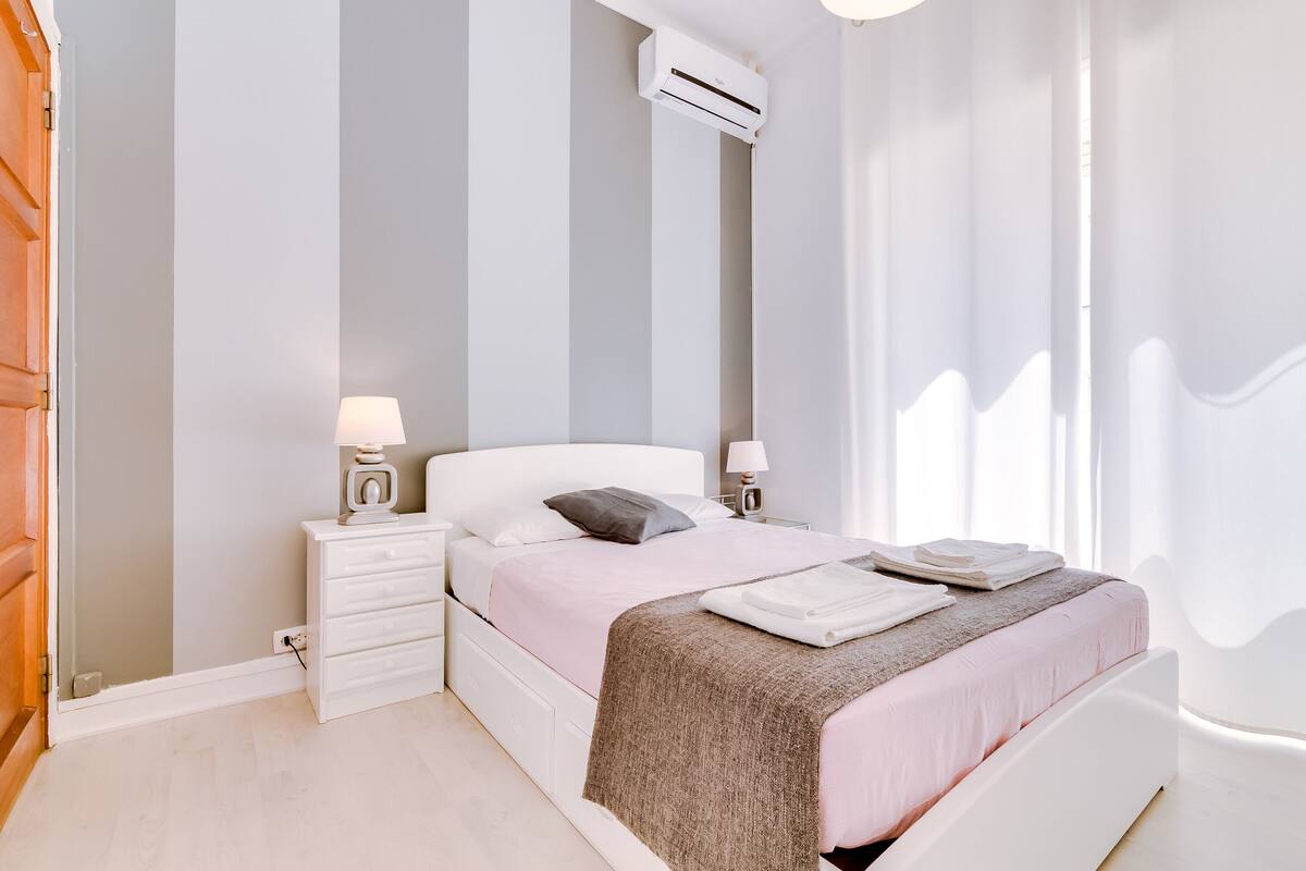 A bedroom with a bed, bedside tables, an air conditioning unit, and curtains.