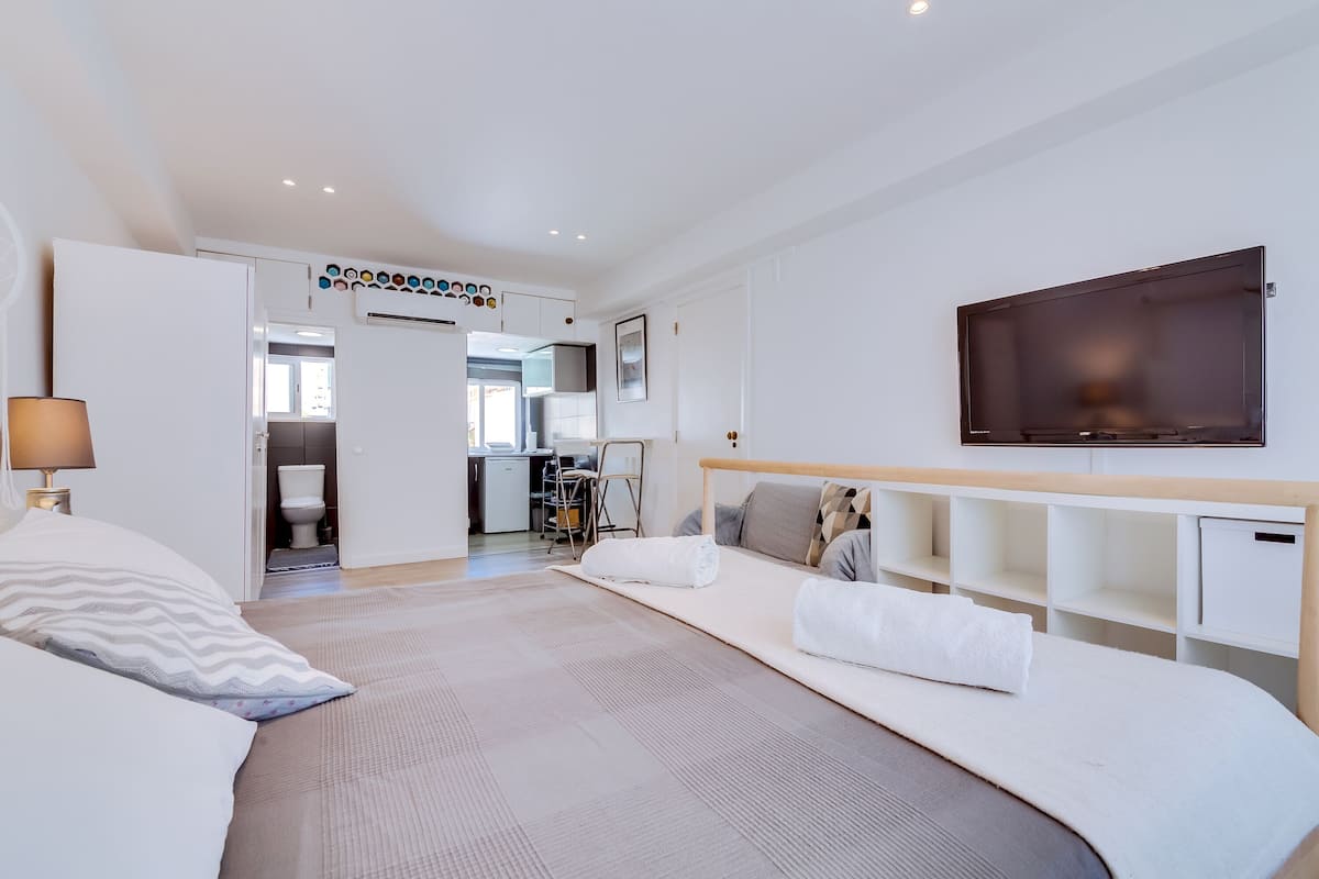 A modern hotel room with a large bed, a TV, and a kitchenette in the background.