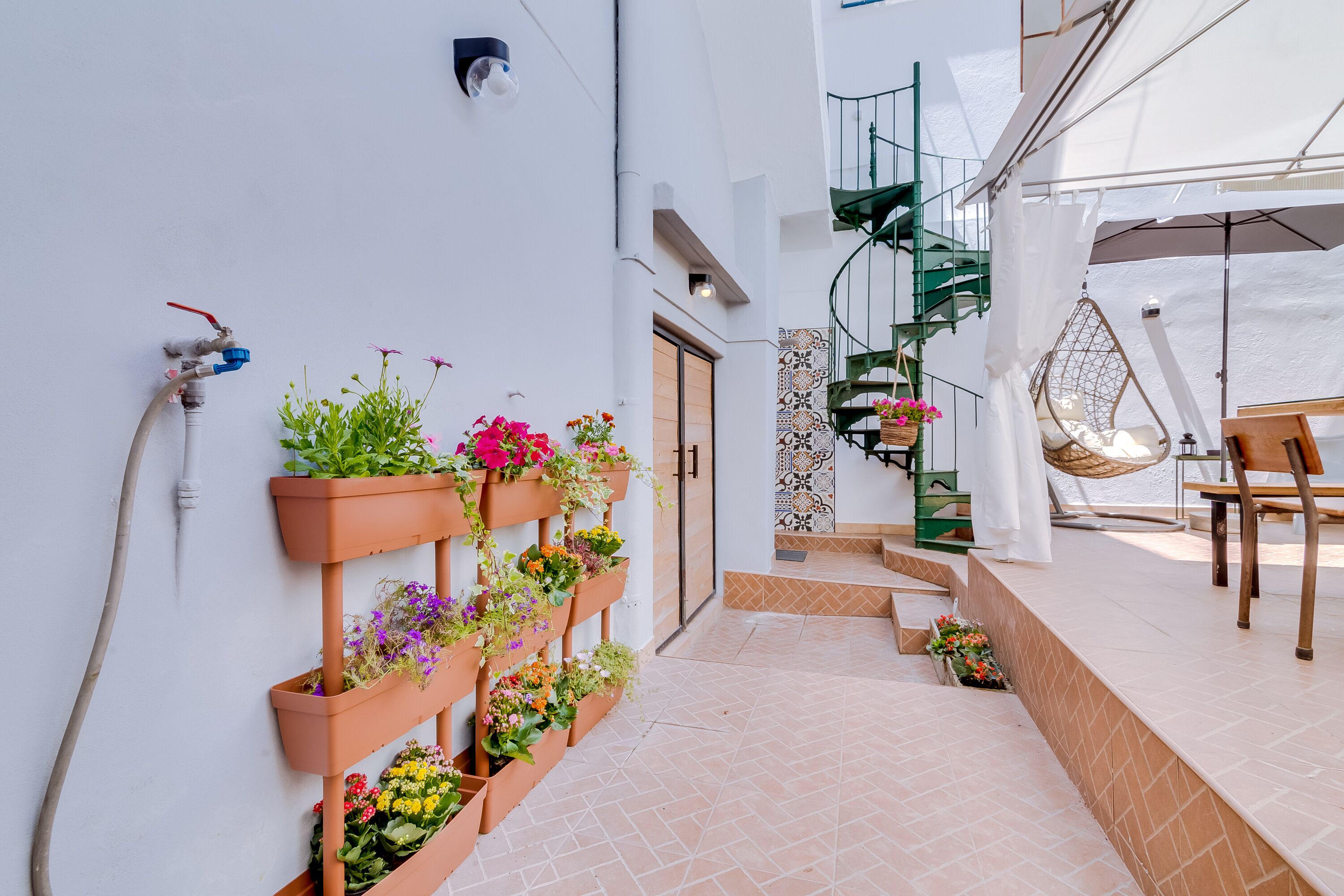 A narrow outdoor corridor with a tiled floor, a wall-mounted water tap, and a series of terracotta planters with various flowers.