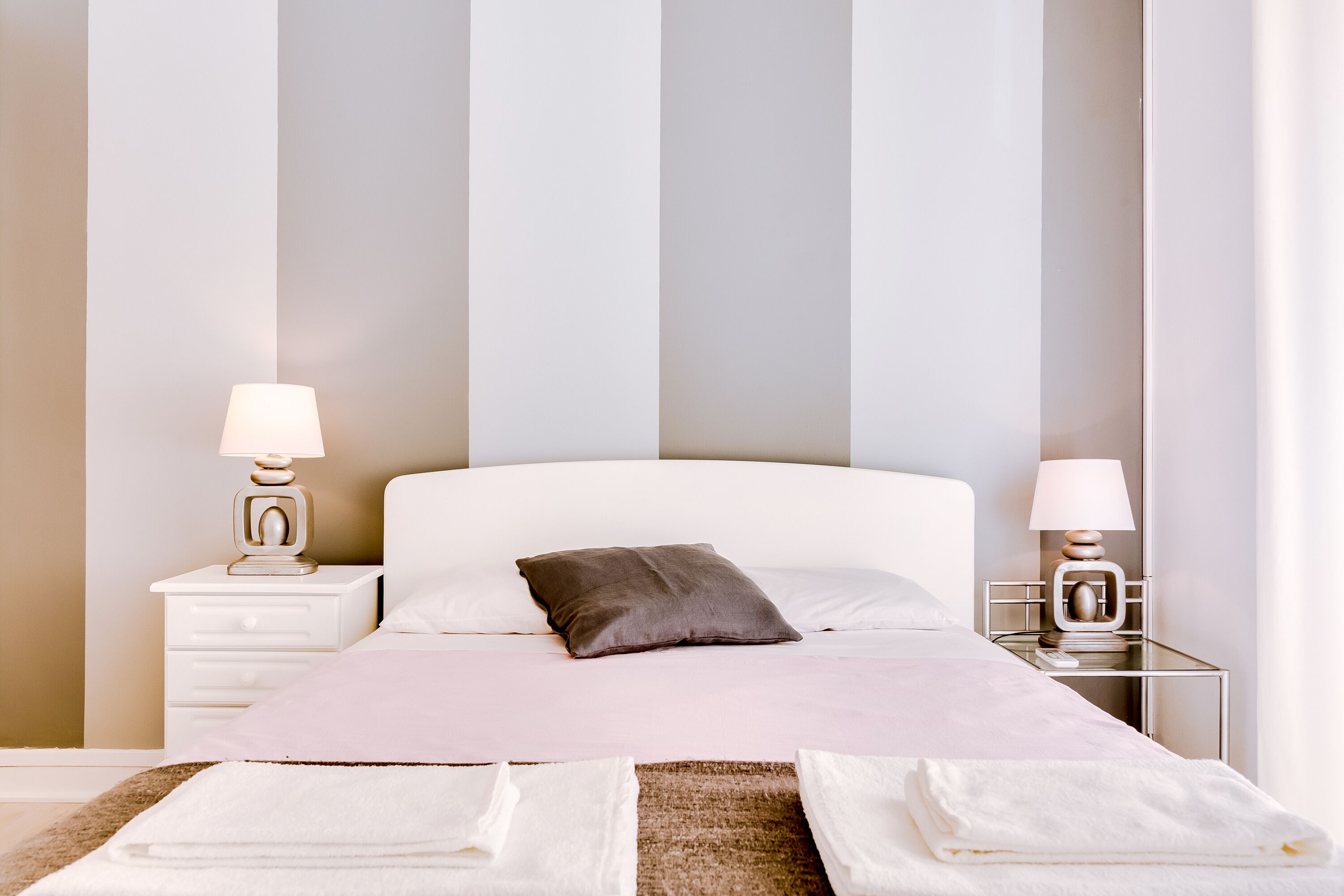 Un lit bien fait, avec une tête de lit blanche, deux tables de chevet avec des lampes et, en arrière-plan, un mur à rayures.
