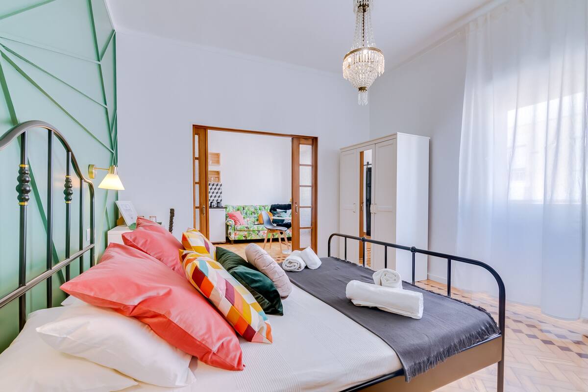 A bedroom with a bed, pillows, a nightstand, a mirror, a wardrobe, and a chandelier.